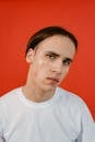 Young man with makeup on his face against a vibrant red background wearing a white t-shirt, looking at the camera.